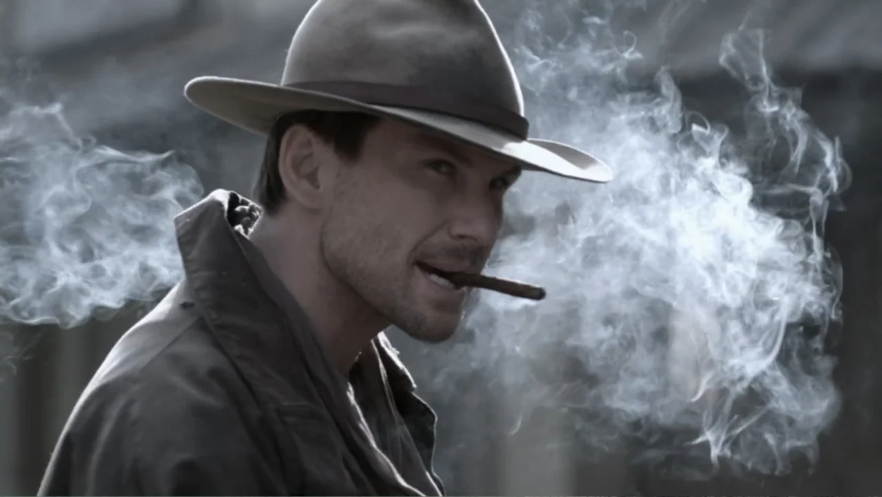 Dawn Rider (2012) movie still: medium — An outdoor setting features a man standing in a diffused light environment. A man wearing a fedora bites on…