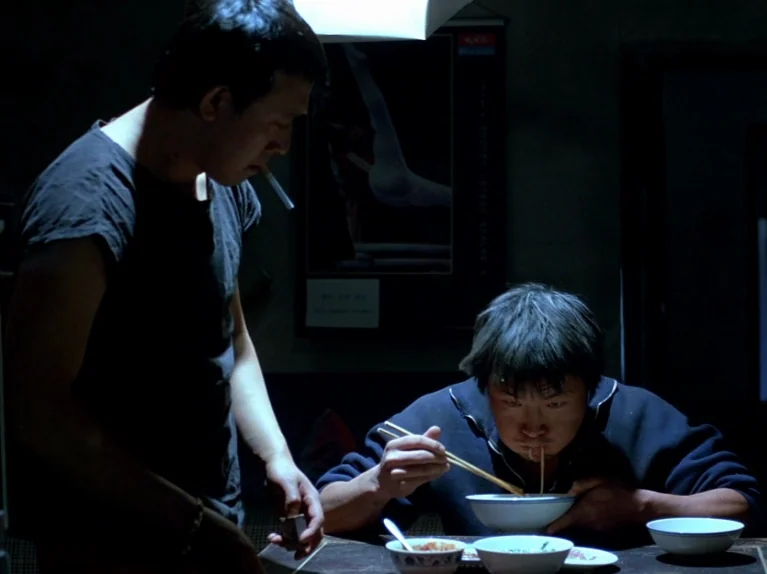Black Snow (1990) movie still: medium — A dimly lit room features a table set with several ceramic bowls of food. One man stands smoking while…