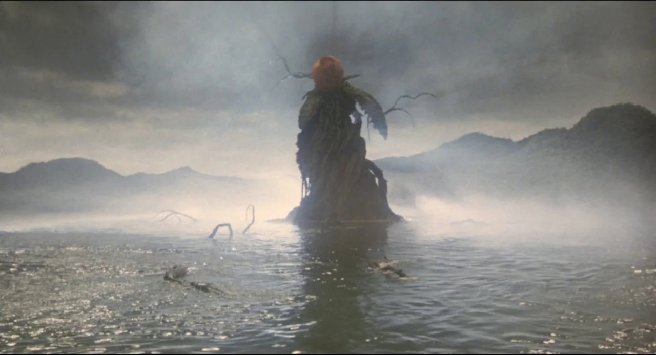 A dark, plant-like entity rises from a calm lake surrounded by misty hills. The environment is shrouded in fog under an overcast sky.