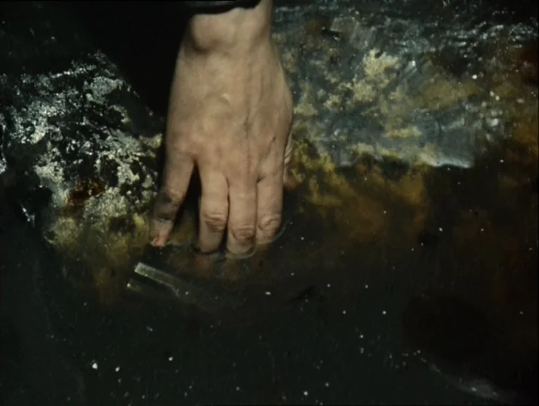 Stalker (1979) movie still: closeup — A dirty, stagnant puddle reflects dim light on a dark, wet ground surface. A human hand reaches down and…
