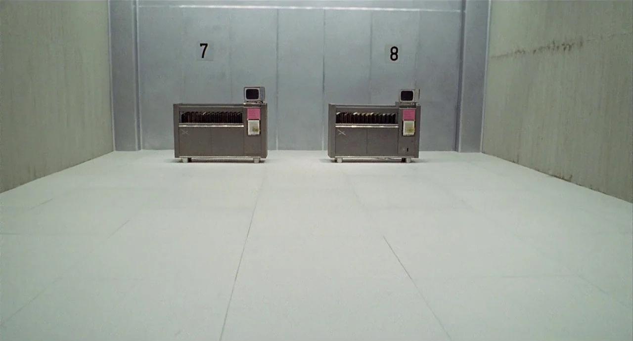 The 10th Victim (1965) movie still: wide — A sterile, minimalist room features stark white floors and clean metal wall panels. Two identical grey…