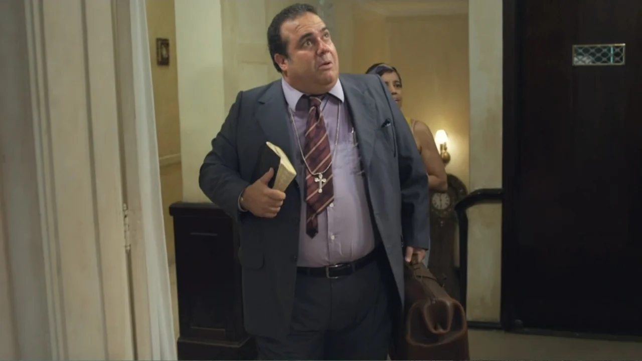 For Sale or Rent (2013) movie still: medium — An interior hallway with beige walls and a dark door frame. A stout man in a suit carries a leather…
