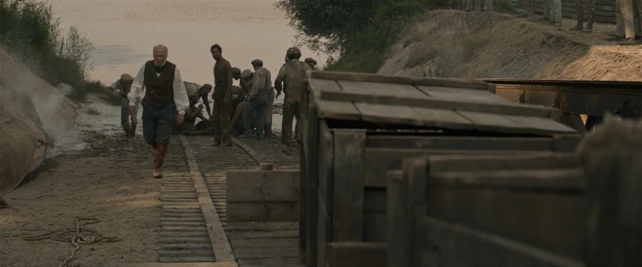 Eiffel (2021) movie still: wide — A rural construction site features railroad tracks running alongside a wide muddy river bank. An older man in…