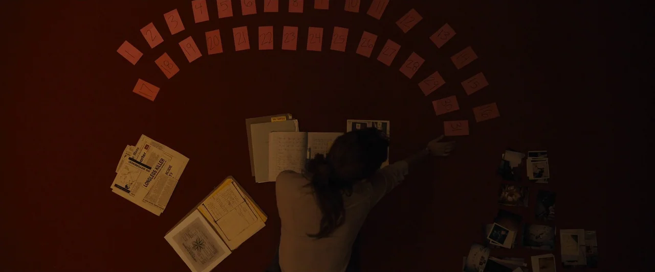 Longlegs (2024) movie still: wide — A person sits on a dark red floor surrounded by documents and photos. The person arranges numbered pink cards in…