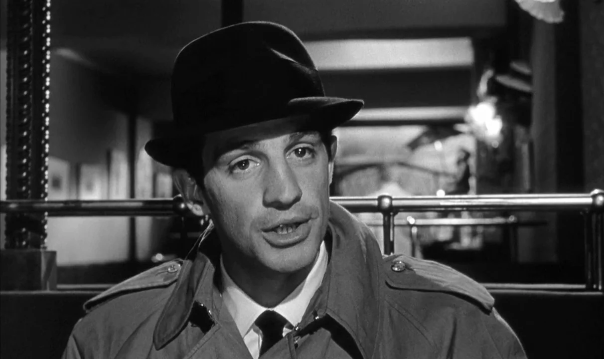 Le Doulos (1962) movie still: closeup — A man sits in a booth at a diner with a metal railing behind him. The subject wears a hat and coat while…