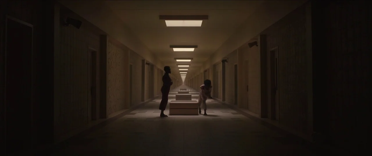 Us (2019) movie still: wide — An exceptionally long, symmetrical hallway features tiled walls and a series of recessed ceiling lights. Two people…