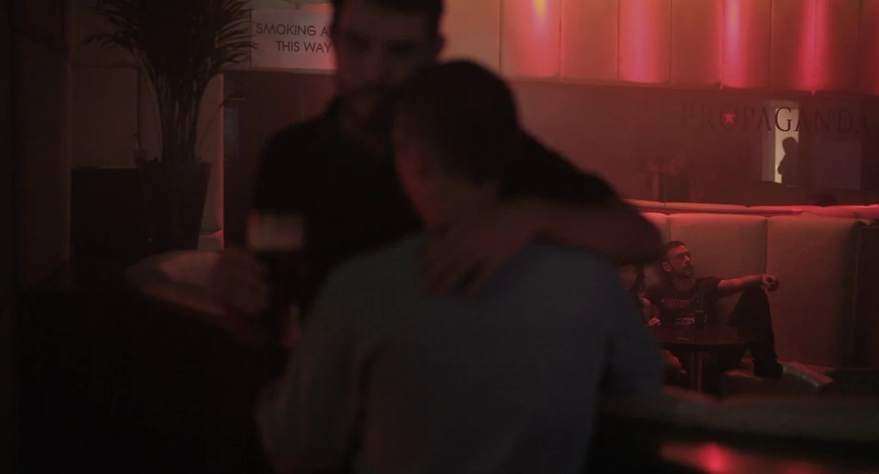 Weekend (2011) movie still: medium — A dimly lit club interior bathed in red light with red-upholstered booth seating. A man in a dark shirt stands…