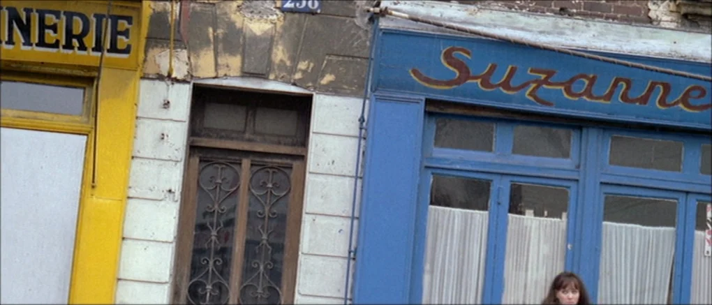 Made in U.S.A. (1966) movie still: medium — A French city street features aging buildings with a yellow storefront and a blue storefront. A young…