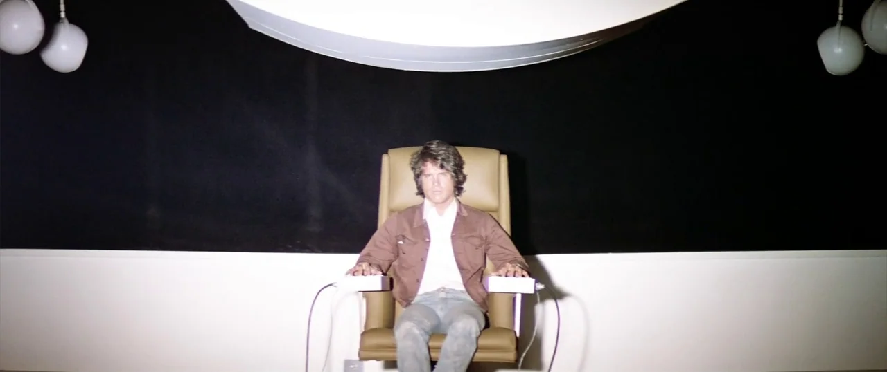 The Parallax View (1974) movie still: medium — A stark room features a high-backed leather chair against a wide black wall panel. A man sits in the…