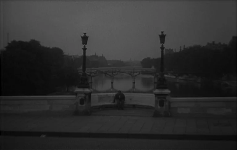 Le Signe du Lion (1962) movie still: wide — A stone bridge over a river in Paris at night features ornate lamp posts. A solitary figure sits on a…