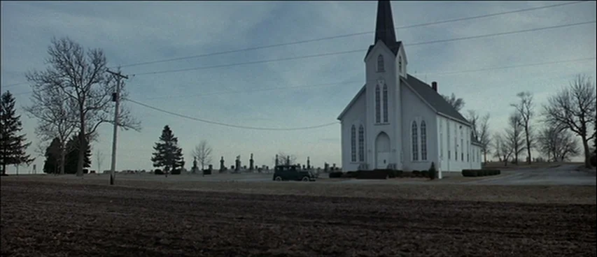 Road To Perdition (2002) movie still: establishing — A rural white church stands next to a cemetery with many headstones under sky. A small vintage…