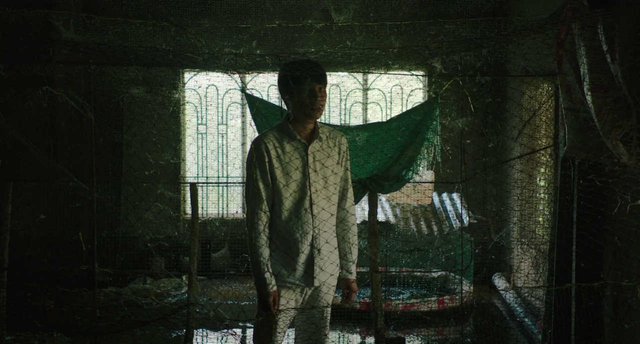 Inside The Yellow Cocoon Shell (2023) movie still: medium — A dimly lit, claustrophobic space is enclosed by rusted wire mesh netting and green…