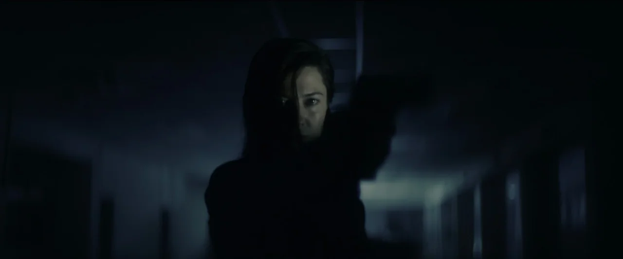 Kate (2021) movie still: medium — A dark hallway with a faint light source illuminating a silhouette. A woman holds a firearm directed toward the…