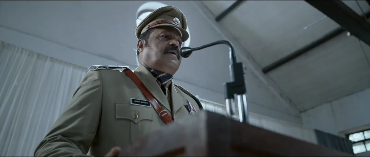 Garudan (2024) movie still: medium — An indoor auditorium features a high vaulted ceiling and light-colored walls with white curtains. A police…