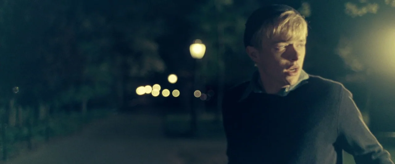 Kill Your Darlings (2013) movie still: medium — A dark night park pathway is illuminated by distant glowing streetlamps in the background. A young…