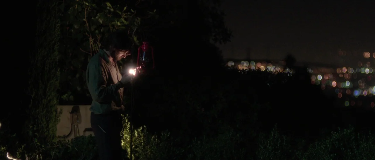 The Invitation (2015) movie still: medium — A man stands outdoors at night on a dark hillside with dense foliage. He holds a lit kerosene lantern,…