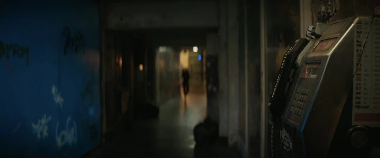 Canary Black (2024) movie still: medium — A dimly lit indoor corridor shows a payphone mounted on a wall. A blurred figure is visible walking in the…