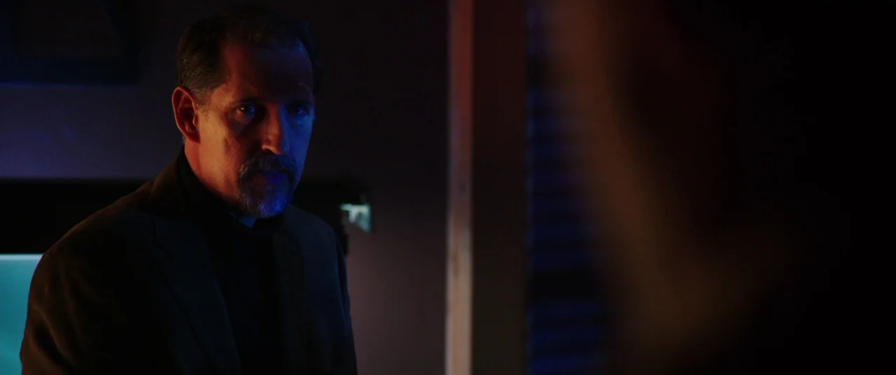The Mind’s Eye (2015) movie still: medium — A dark room with minimal light reflecting on surfaces. A man in a brown blazer glances toward an unseen…