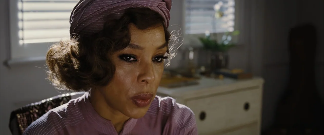 Death on the Nile (2022) movie still: closeup — A woman sits in a room with white wooden furniture and window shutters. The woman is positioned in…