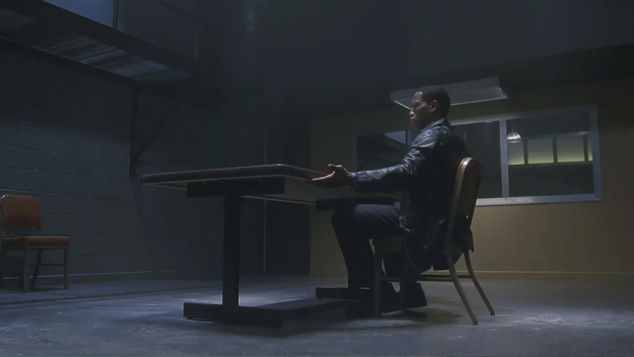 The Art of War (2000) movie still: medium — A dimly lit room features a metal table centered on a concrete floor. A man sits alone at the table with…