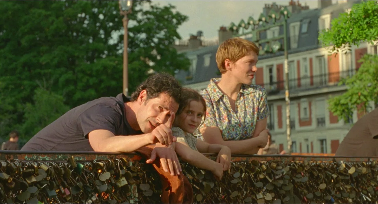 One Fine Morning (2022) movie still: medium — An outdoor urban bridge is covered in numerous metal padlocks along the iron railing. A man, a young…