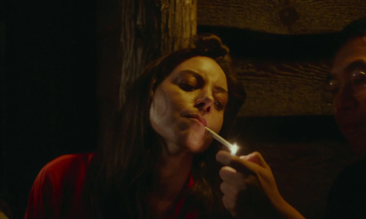 Black Bear (2020) movie still: closeup — A dark wooden room with logs visible in the background. A woman in a red shirt leans toward a lighter held…