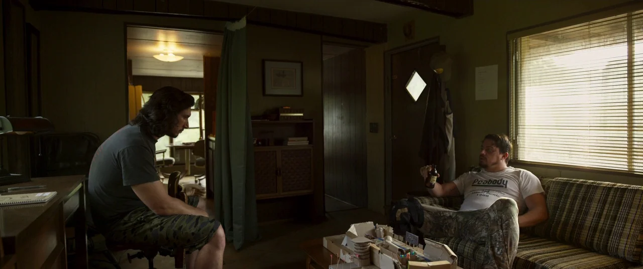 Logan Lucky (2017) movie still: wide — A dimly lit room features wood-paneled walls and a striped couch with blinds on window. Two men sit in the…