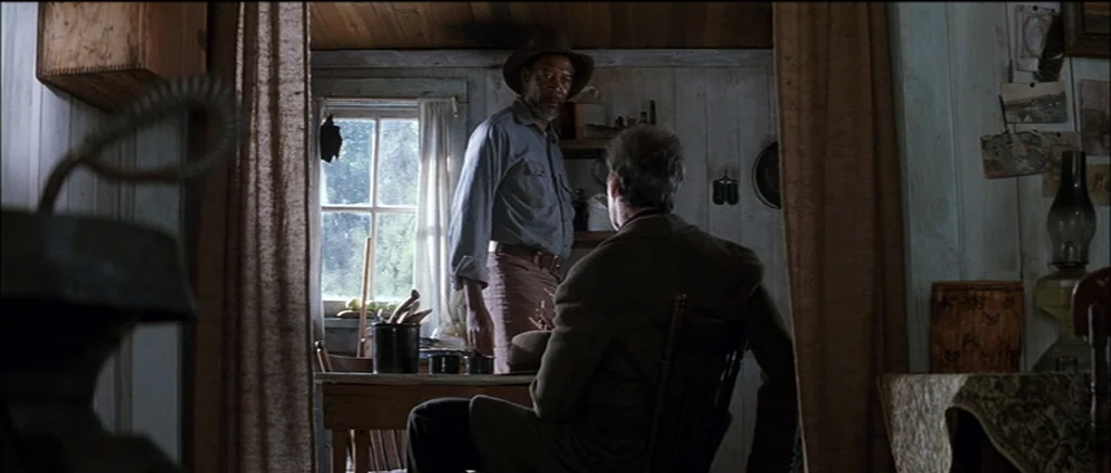 Unforgiven (1992) movie still: medium — A small rustic cabin interior has wooden plank walls and a simple dining area. A standing man in a hat…