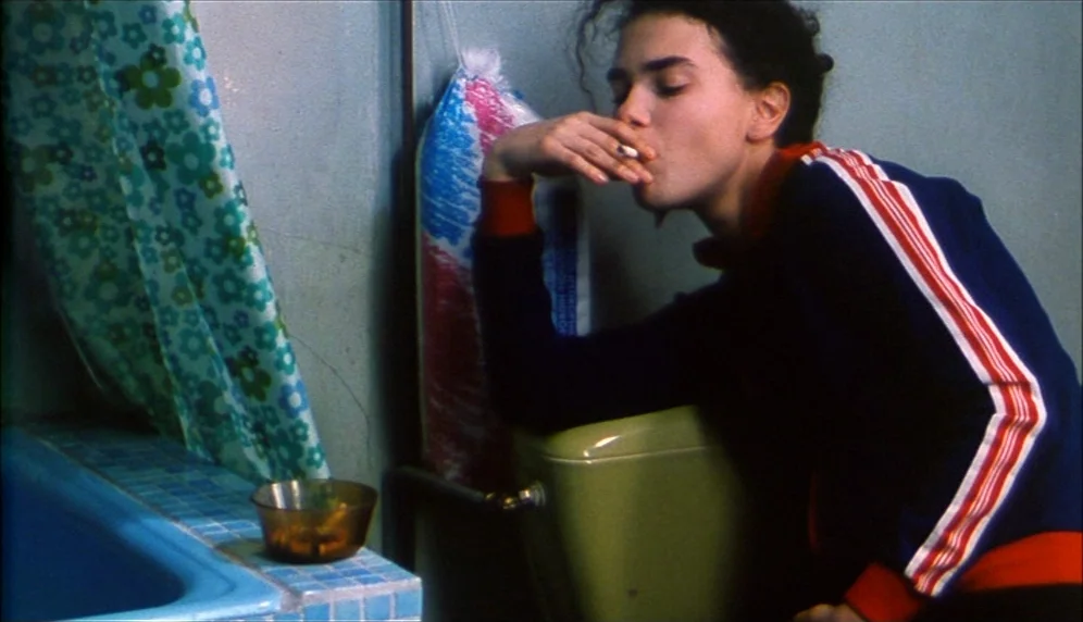Nenette et Boni (1996) movie still: medium — A small bathroom features a green toilet tank and a blue tiled edge. A young woman leans against the…