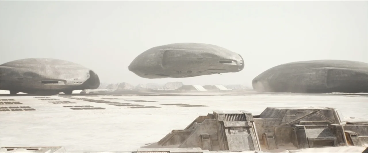 Dune (2021) movie still: wide — A flat, sun-bleached desert basin features large, metallic, disc-shaped structures resting on the ground. Several…