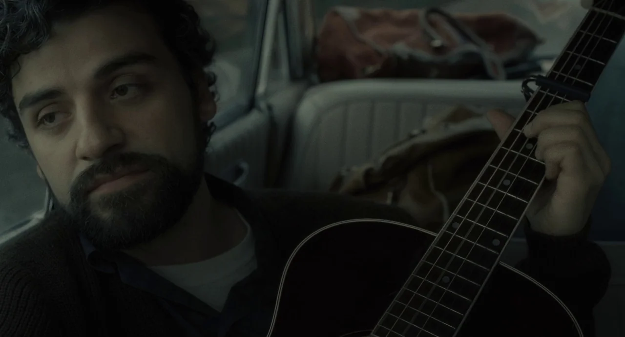 Inside Llewyn Davis (2013) movie still: closeup — A man sits inside the backseat of a vintage car with tan upholstery. He holds an acoustic guitar…