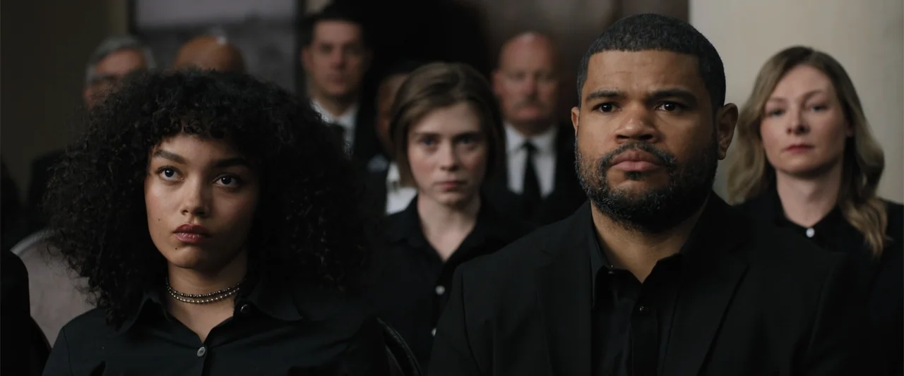 Trap House (2025) movie still: medium — A group of people sit in rows within a pale, minimalist interior space. The subjects face forward in formal…