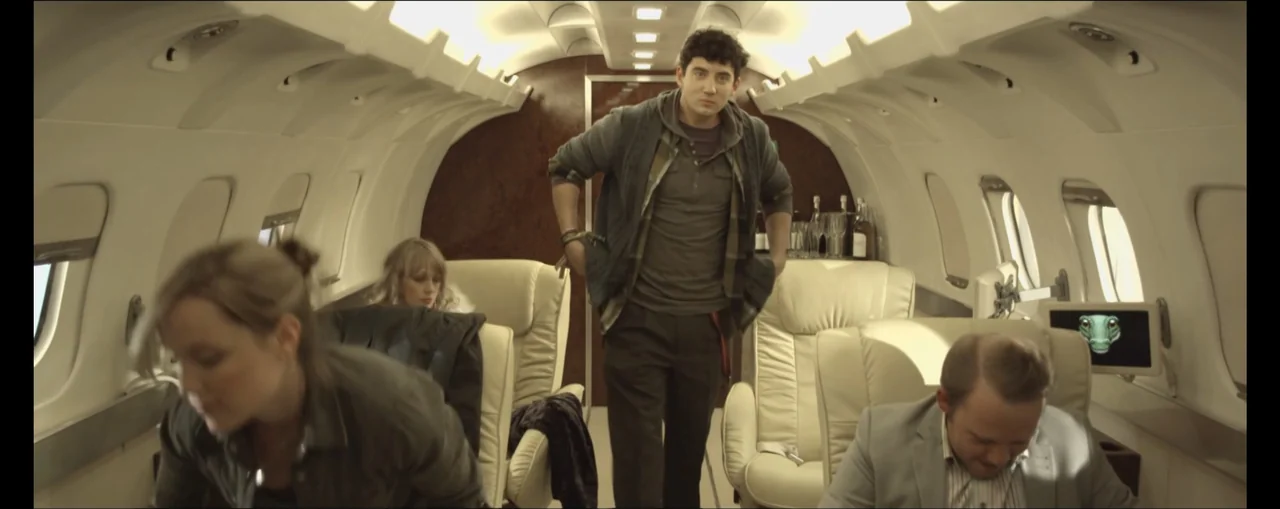 Turbulence (2025) movie still: medium — The interior of a private jet features light-colored leather seats and dark wood paneling. A man stands in…