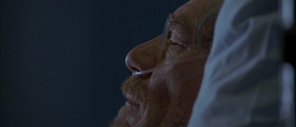 Apt Pupil (1998) movie still: closeup — A dimly lit hospital room at night with dark blue shadows surrounding the bed. An elderly man with a…