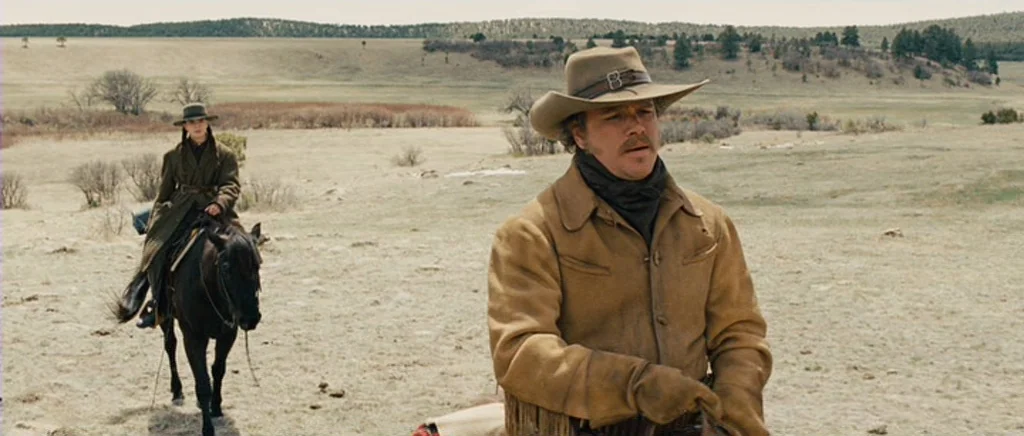 True Grit (2010) movie still: wide — An open landscape with dry vegetation and distant trees under an overcast sky. Two riders move across the…