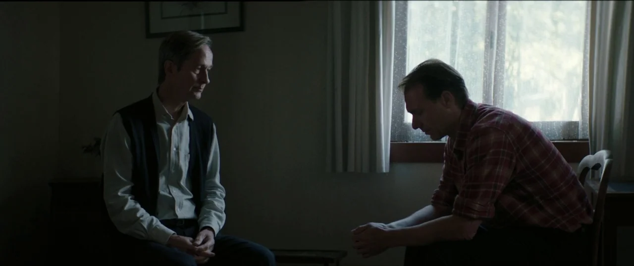 Good Favour (2017) movie still: medium — A dimly lit interior room with a window covered by thin white curtains. Two men sit on wooden chairs…