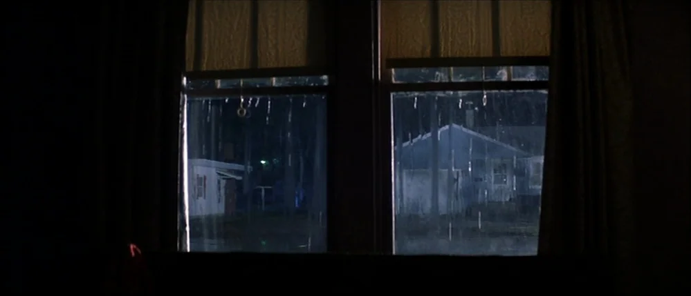 The Straight Story (1999) movie still: wide — A dark, interior view looking out through a window at a rainy night scene. Rain streaks down the glass…