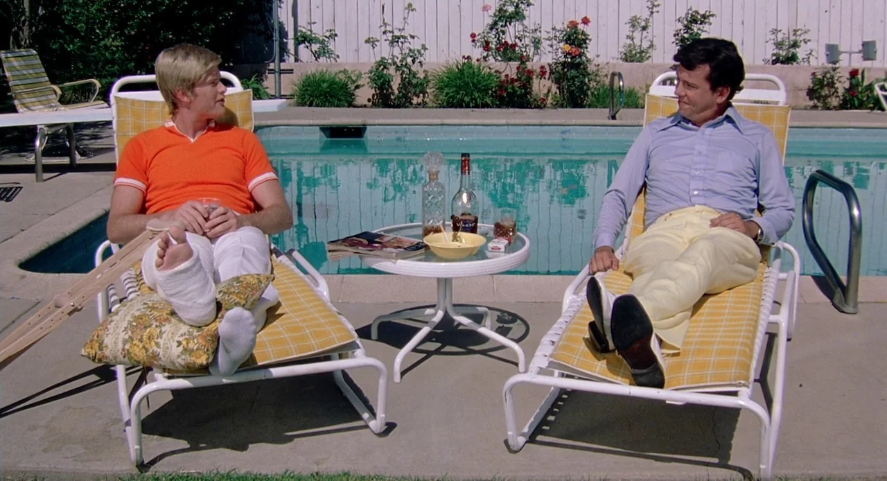 Viva (2007) movie still: wide — A sunny suburban swimming pool deck with white fences and rose bushes behind. Two men sit on matching yellow plaid…