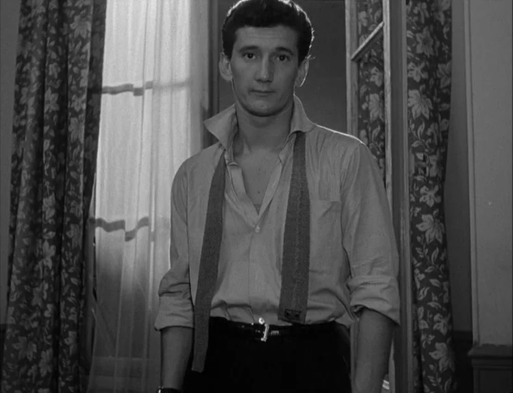 Bob le Flambeur (1956) movie still: medium — A domestic space with floral print curtains framing a window and light walls. A young man stands in the…