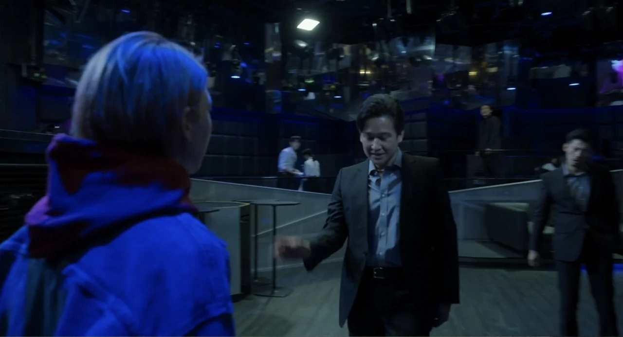 Knuckle Girl (2023) movie still: medium — A dimly lit club interior with dark walls and scattered tables under blue lighting. A man in a suit stands…