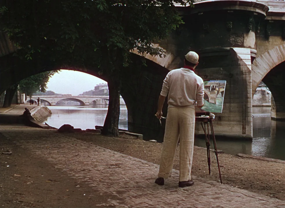 An American In Paris (1951) movie still: fullbody — An artist stands on a dirt path under the arched stone bridge of a river. He is painting a…