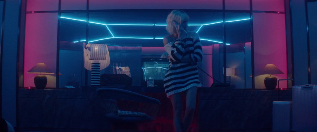 Atomic Blonde (2017) movie still: fullbody — A hotel room at night features neon blue light strips against a dark wall. A woman in a striped sweater…