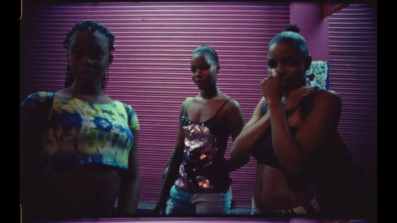 Black Mother (2018) movie still: medium — Three young women stand against a closed corrugated purple metal security shutter at night. The subjects…