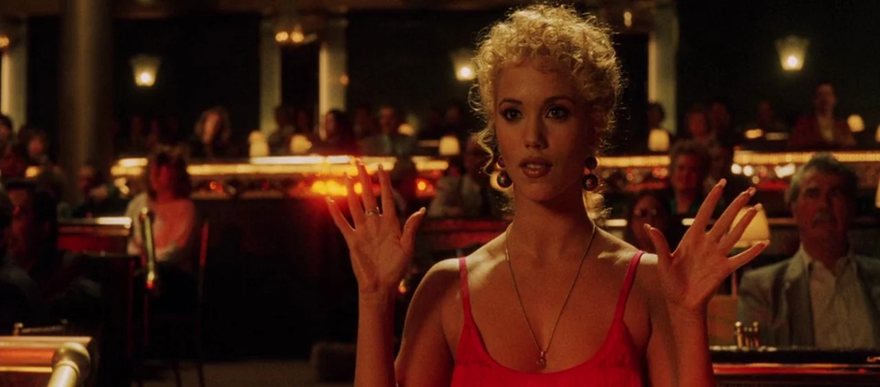 Showgirls (1995) movie still: medium — A dimly lit nightclub interior with a crowd sitting at tables in the blurred background. A woman in a red…