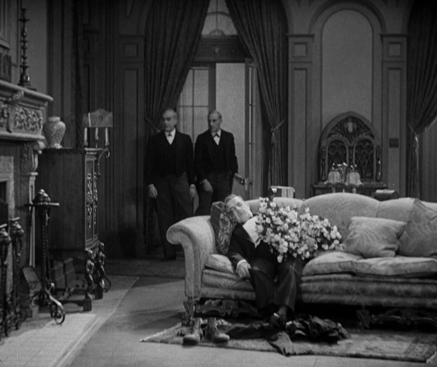City Lights (1931) movie still: wide — An opulent parlor with heavy drapes and wood-paneled walls features an ornate sofa. A man sleeps on the couch…