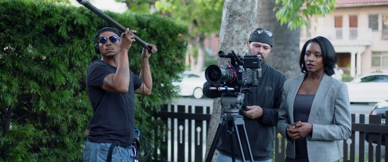 Blindfire (2020) movie still: medium — A film crew works on a tree-lined residential street with houses in the background. A camera operator and a…