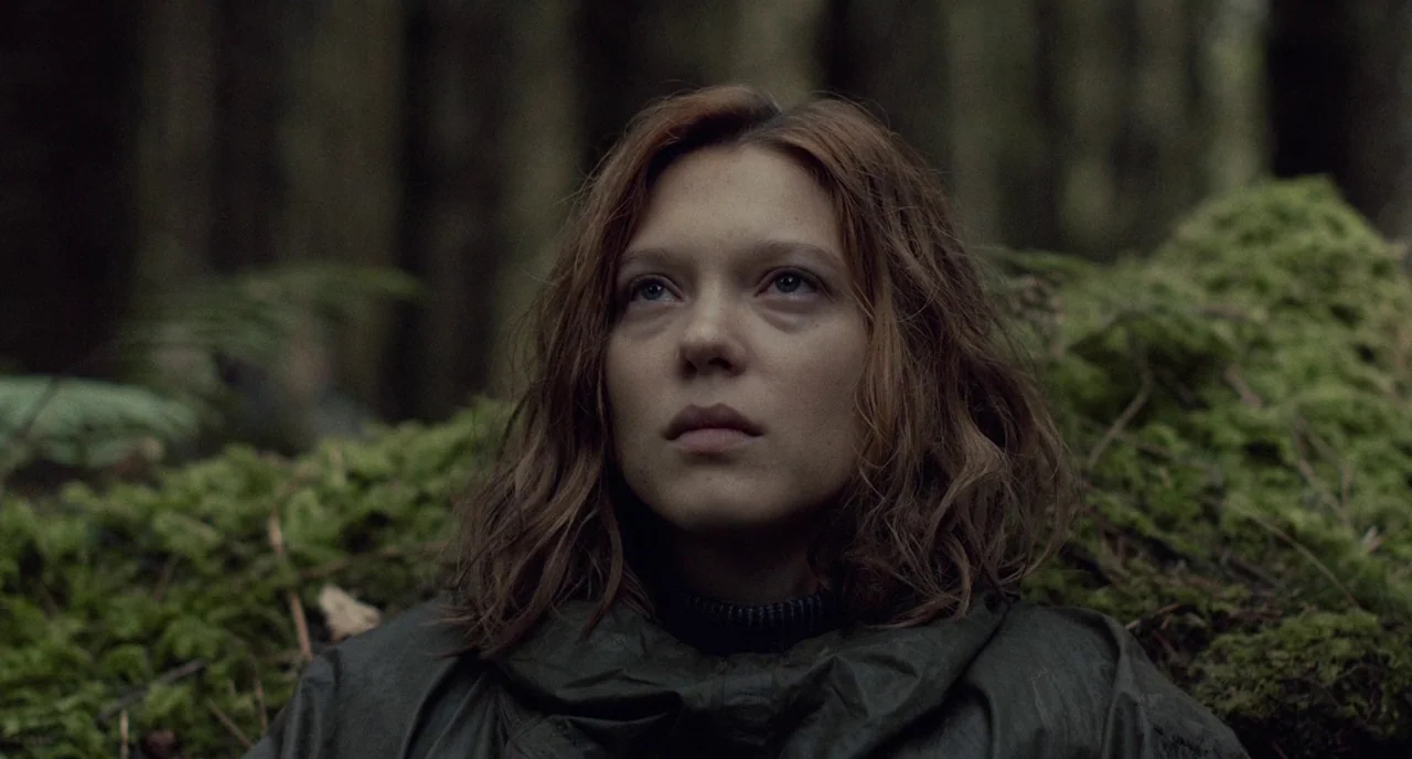 The Lobster (2015) movie still: closeup — A dense forest environment with thick moss growing on a fallen log. A young woman looks toward the sky…