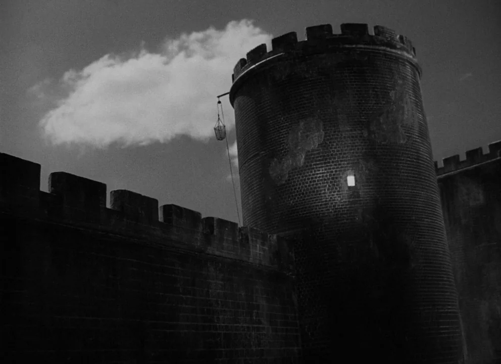 Othello (1952) movie still: establishing — A dark stone fortification wall features a tall, cylindrical tower with battlements at the top. A single,…
