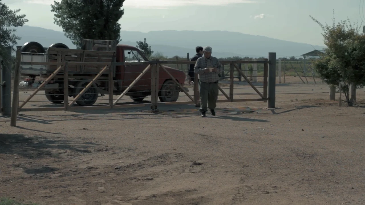 Still Life (2013) movie still: wide — A rural dirt property features a wooden gate and a parked red pickup truck. An older man walks through the…
