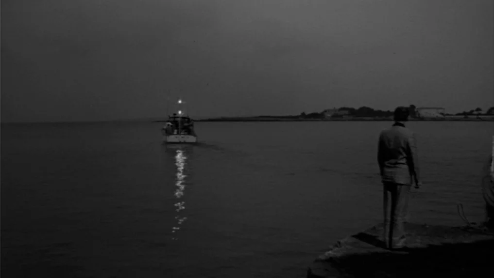 Under The Cherry Moon (1986) movie still: wide — A man stands at the edge of a dark stone pier at night. A small boat with illuminated lights…